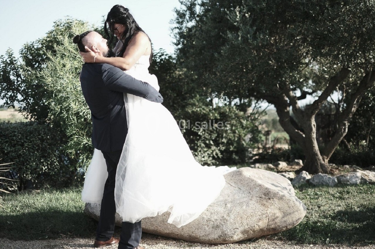 Un couple en tenue de mariage, l'homme portant la femme, dans un jardin avec des arbres et un gros rocher.