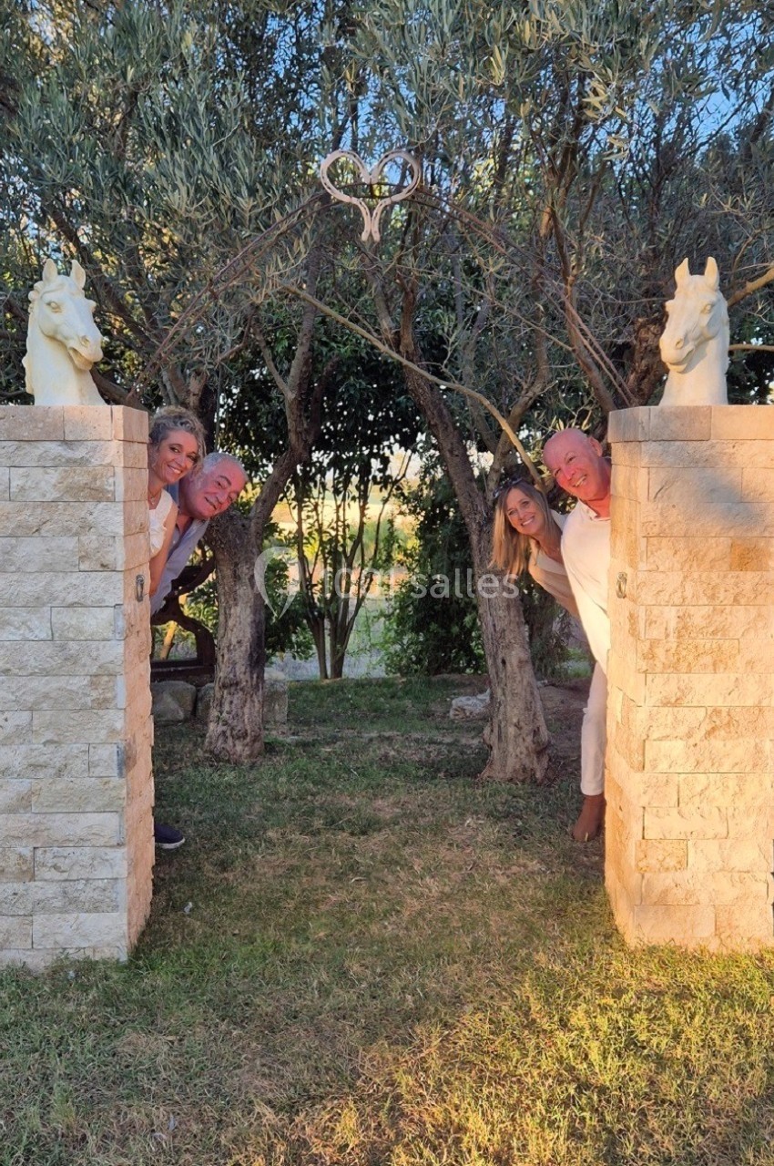 Quatre personnes souriantes se cachent derrière deux piliers en pierre surmontés de statues de chevaux, dans un jardin au…