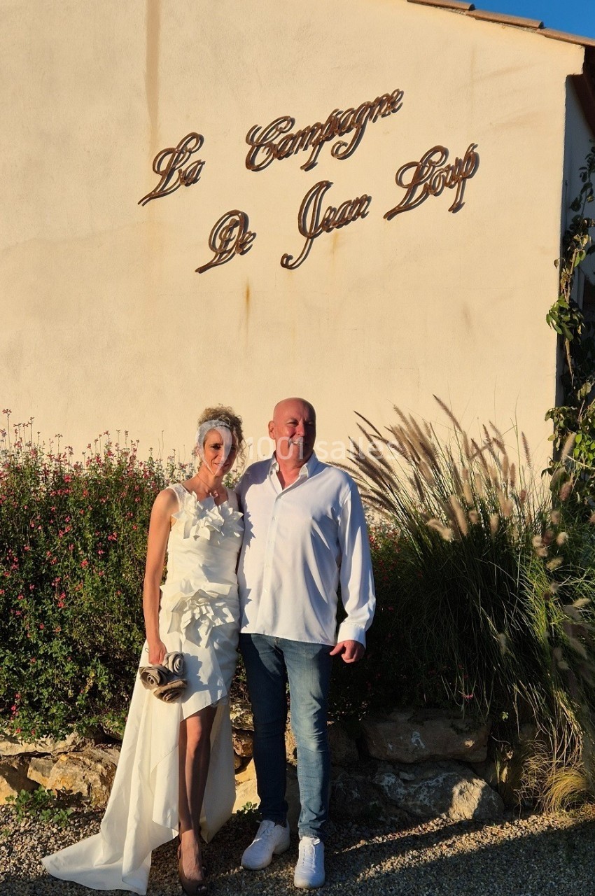 Un couple pose devant un mur beige avec l'inscription ’La Campagne de Jean Loup’, entouré de végétation.