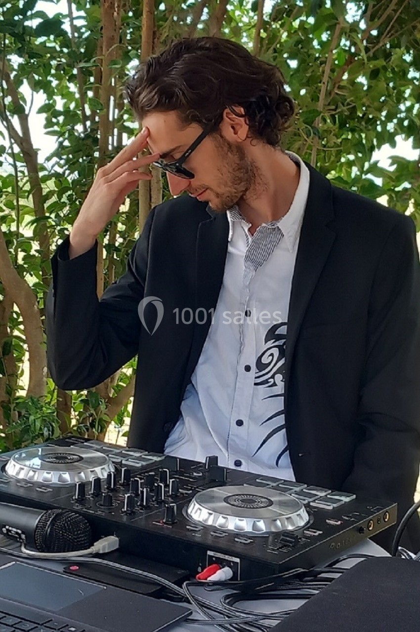 Un DJ en costume noir ajuste son casque devant une table de mixage, en extérieur avec des arbres en arrière-plan.