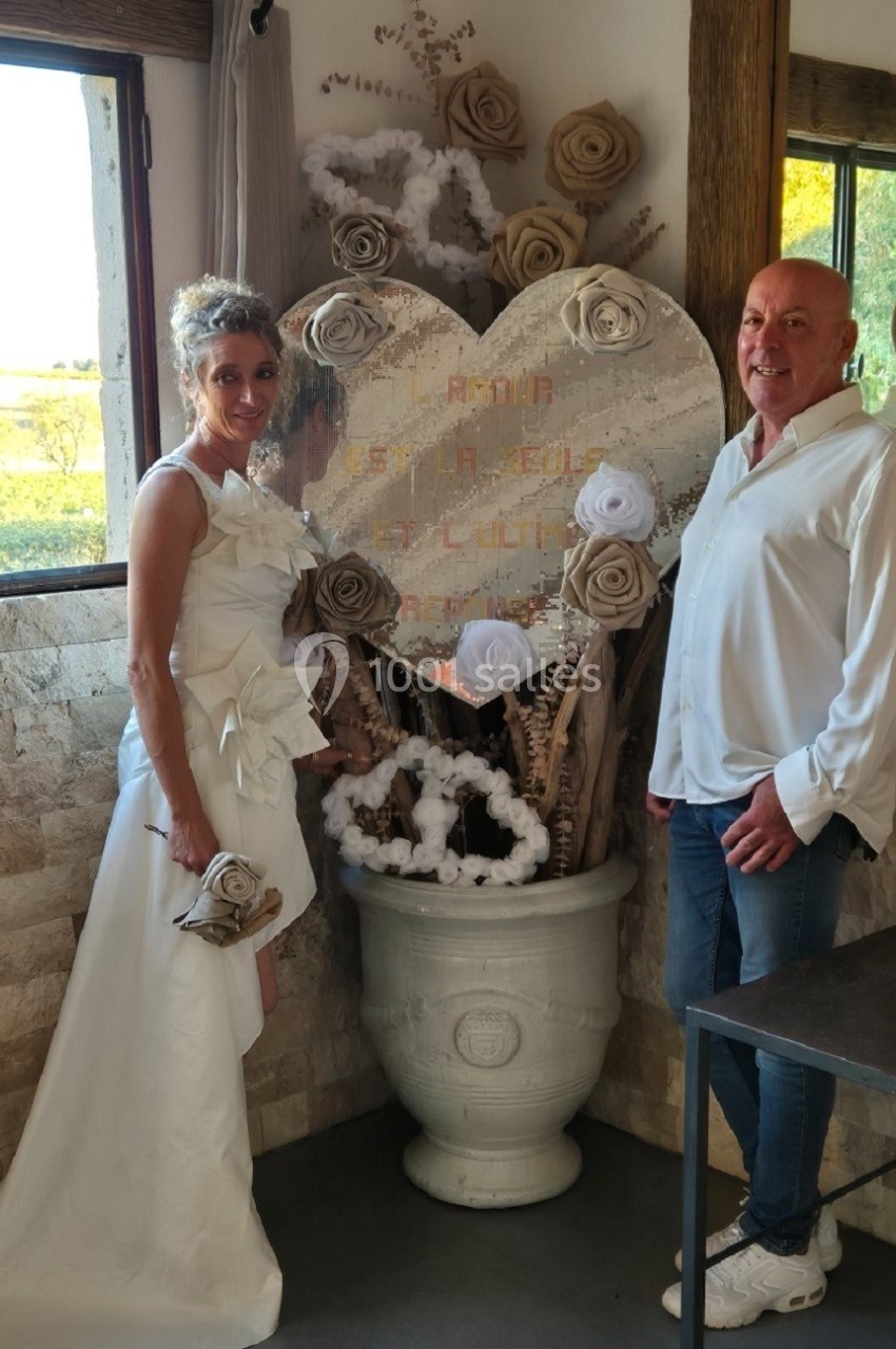 Une femme en robe blanche et un homme en chemise posent près d'une décoration en forme de cœur dans une salle lumineuse.