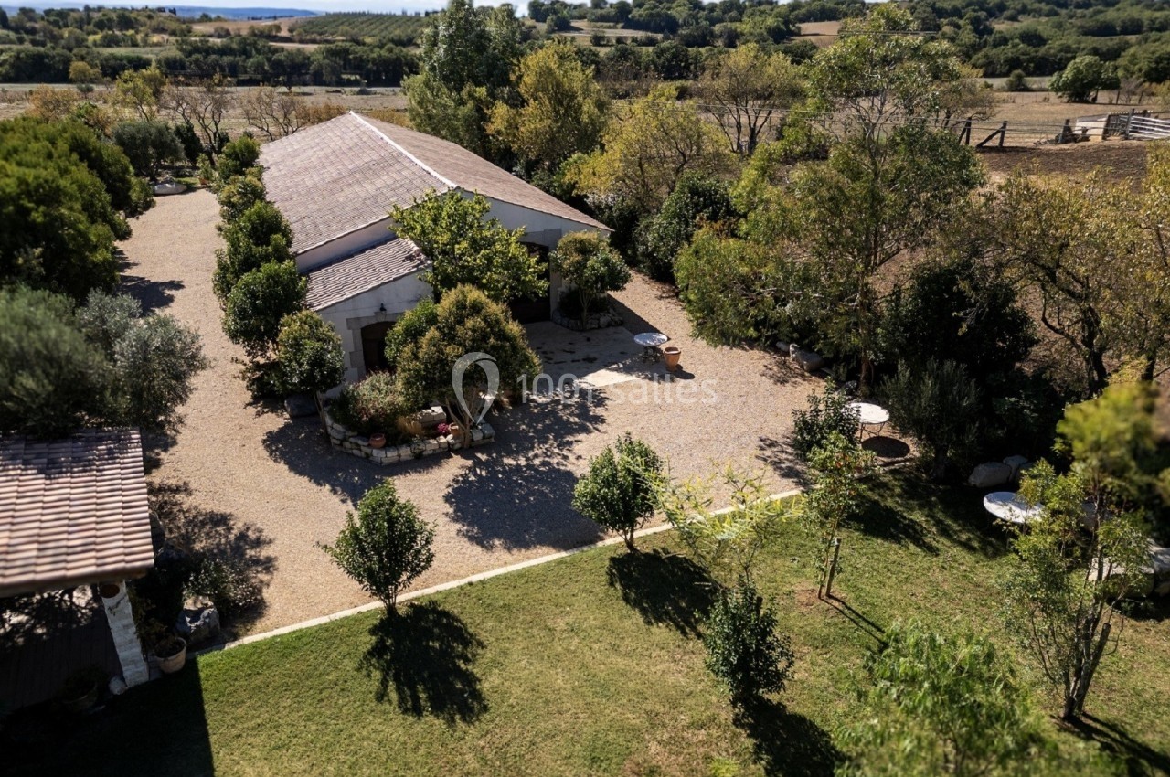 Vue aérienne d'une maison en pierre entourée d'arbres, avec une cour gravillonnée et un paysage rural en arrière-plan.