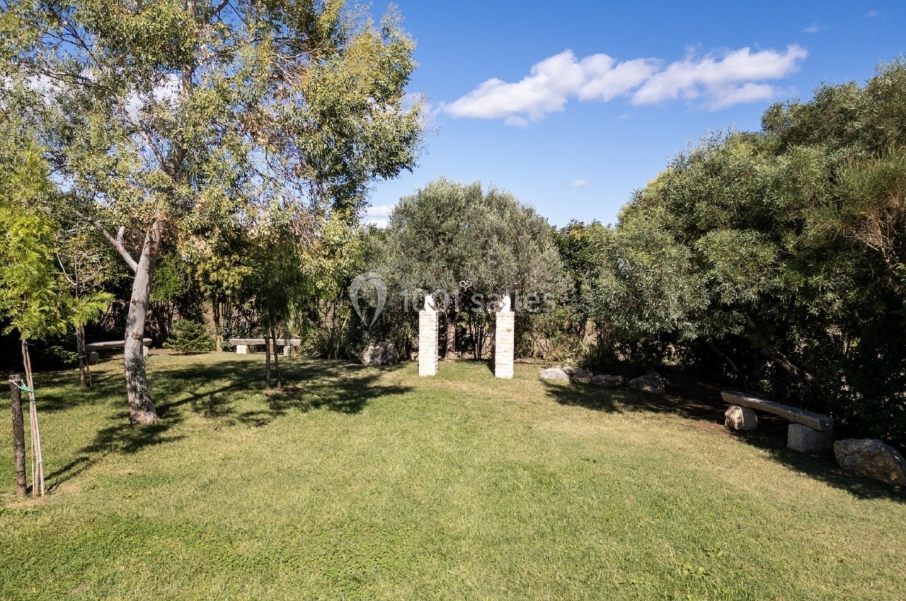 Pelouse verdoyante avec arbres, bancs en pierre et deux piliers en pierre sous un ciel bleu dégagé.