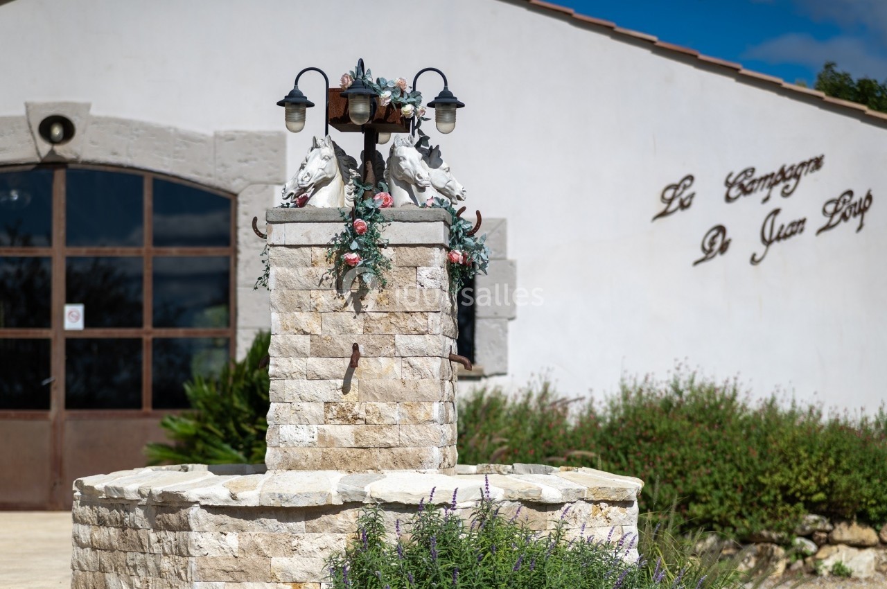 Fontaine en pierre ornée de sculptures de chevaux et de fleurs, devant un bâtiment blanc avec une inscription murale.