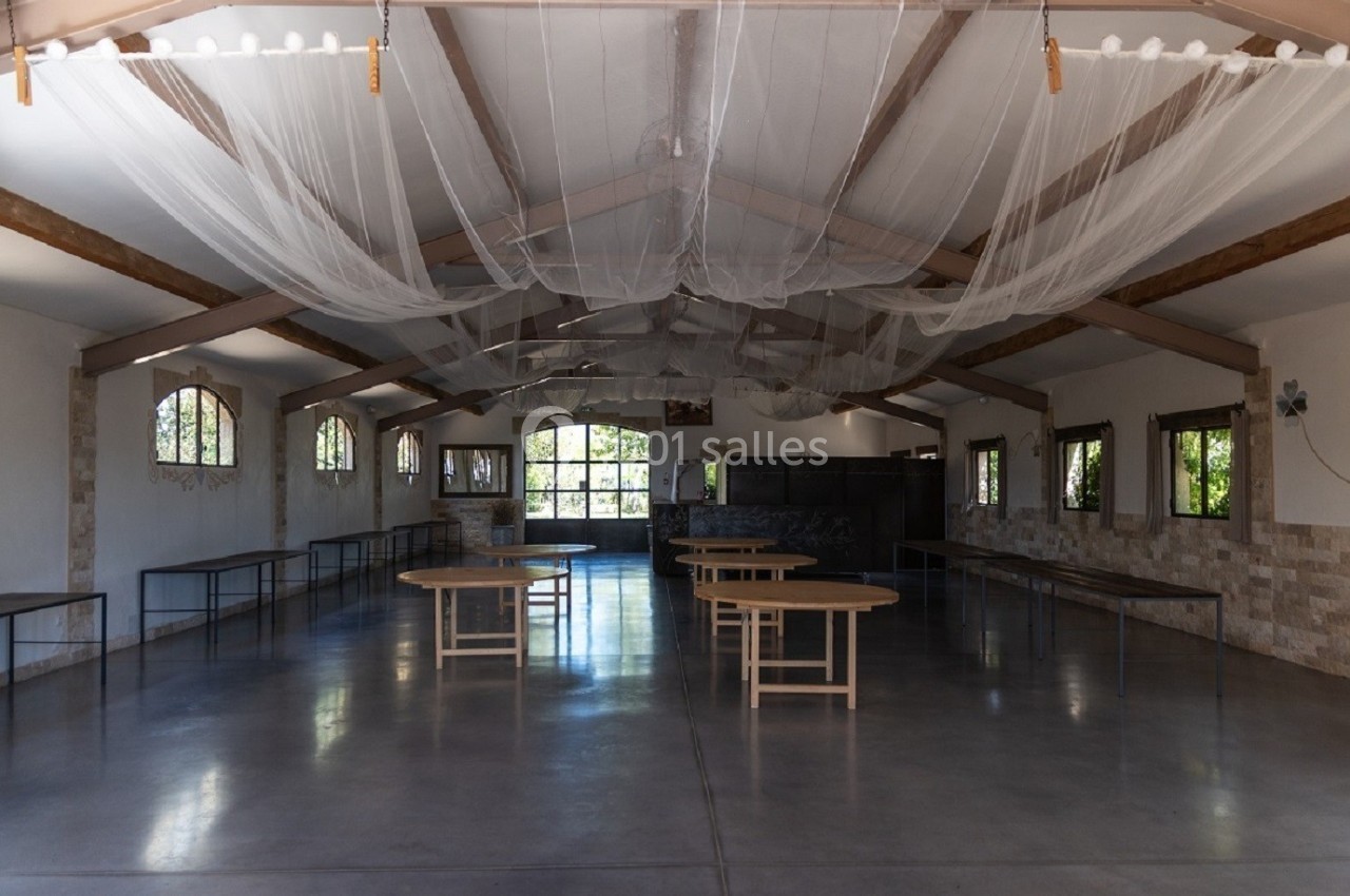 Grande salle lumineuse avec tables rondes en bois, sol en béton poli, poutres apparentes et filets suspendus au plafond.
