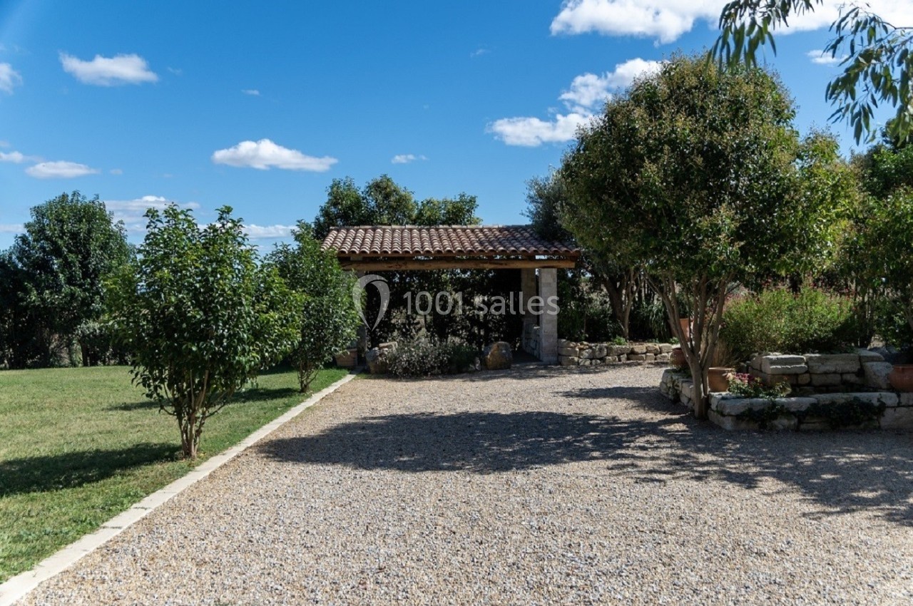 Allée en gravier menant à un abri en pierre avec toit en tuiles, entouré de végétation et d'arbres sous un ciel bleu.