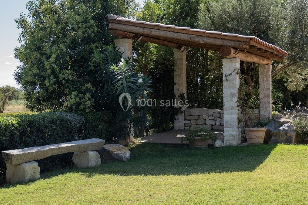 Abri en pierre et bois entouré de végétation, avec un banc en pierre sur une pelouse ensoleillée.