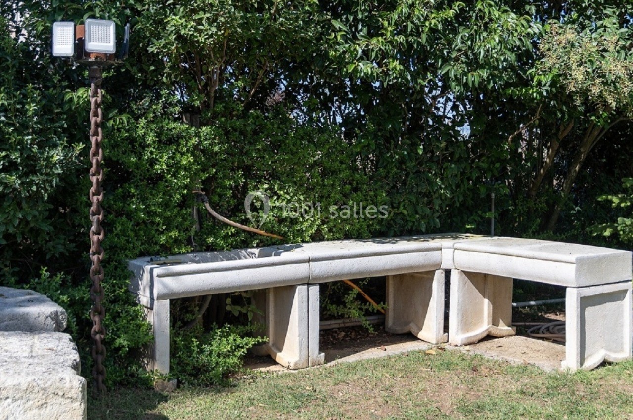 Banc en béton courbé installé dans un jardin, entouré de végétation et éclairé par un projecteur sur pied.