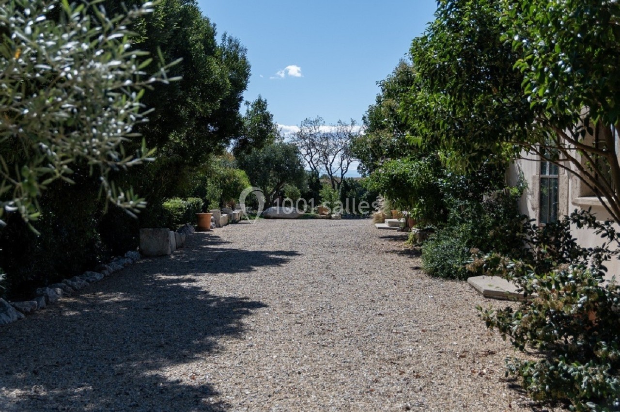 Allée en gravier bordée de végétation méditerranéenne et d'arbres, avec une maison visible sur la droite.