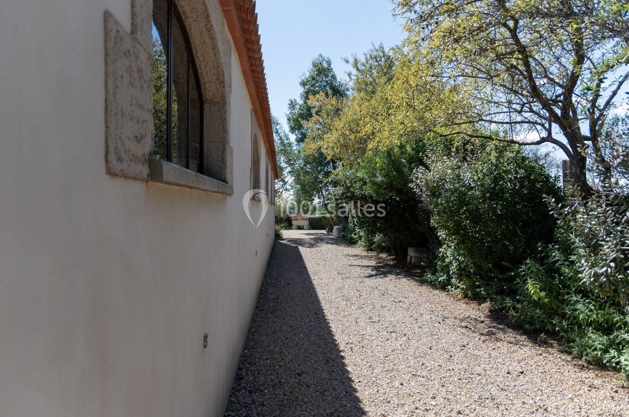 Allée en gravier longeant un bâtiment avec des fenêtres en arc, bordée d'arbustes et d'arbres sous un ciel dégagé.