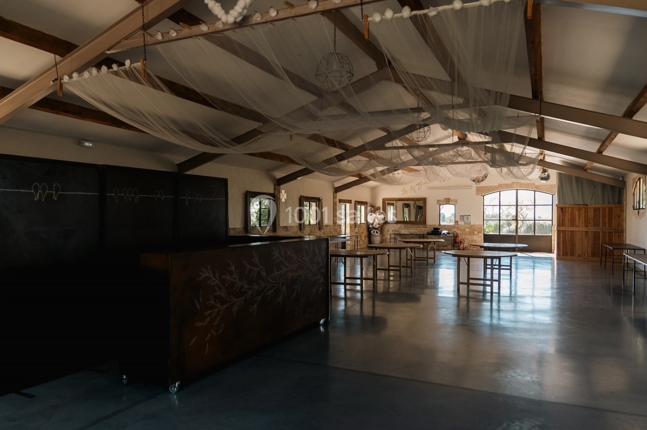 Salle spacieuse avec poutres apparentes, tables et décorations suspendues, éclairée par de grandes fenêtres.