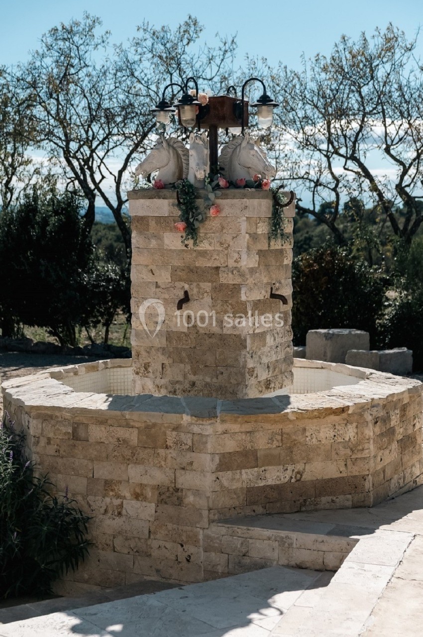 Fontaine en pierre rustique avec deux robinets, ornée de fleurs et lanternes, entourée de végétation et d'arbres.