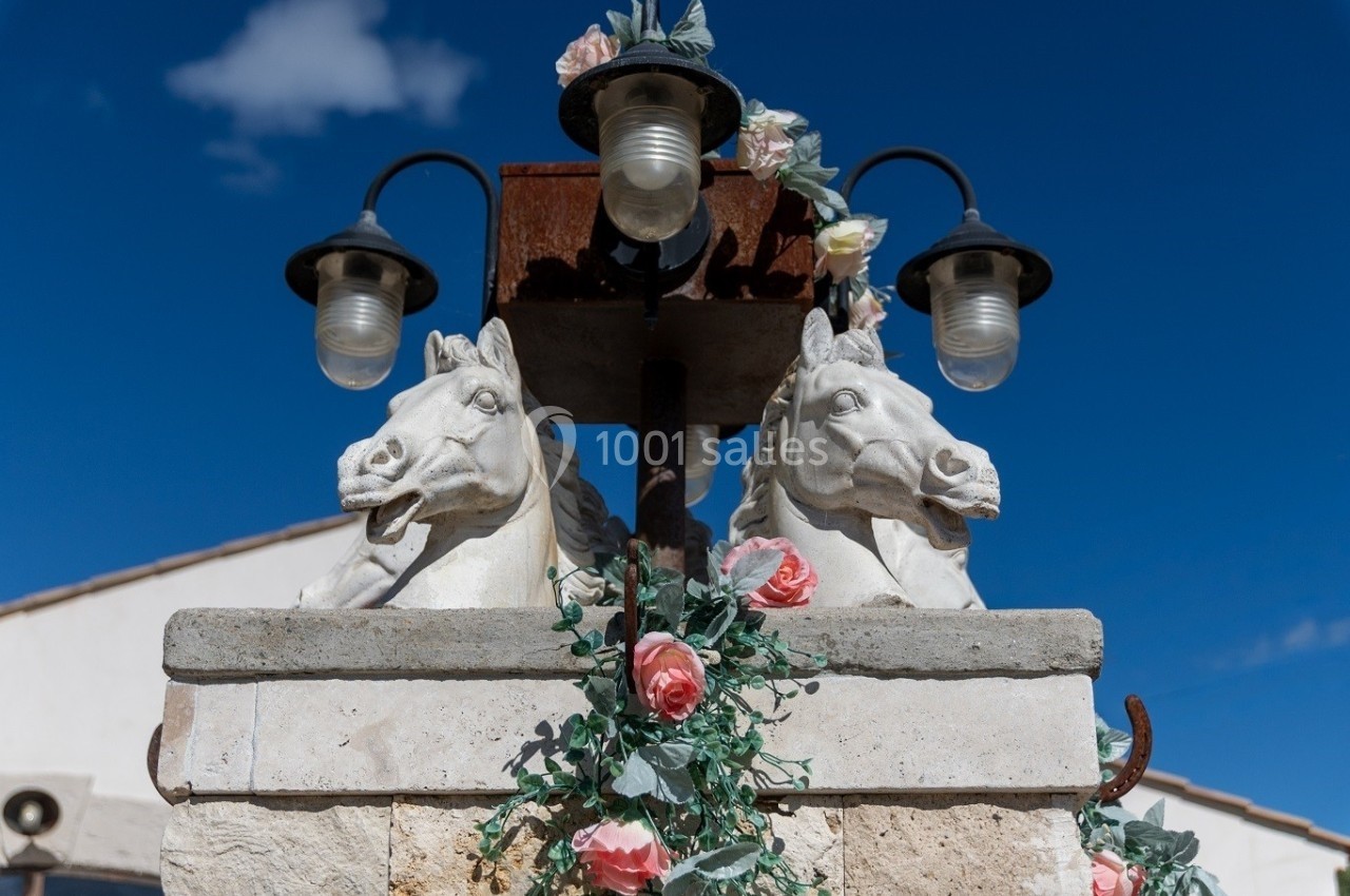 Statues de têtes de chevaux ornées de guirlandes de fleurs, surmontées de lampes, devant un ciel bleu dégagé.