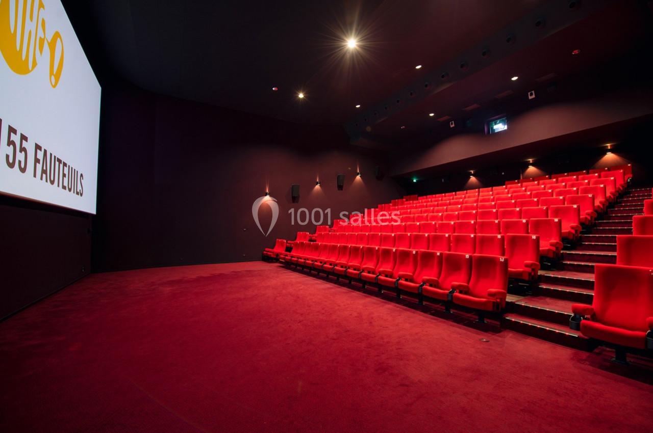 Salle de cinéma vide avec des rangées de fauteuils rouges et un écran blanc allumé à gauche.
