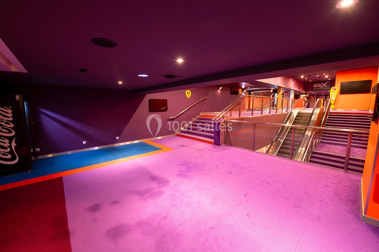 Escalier et escalator dans un hall de cinéma aux couleurs vives avec moquette violette et affichages lumineux.