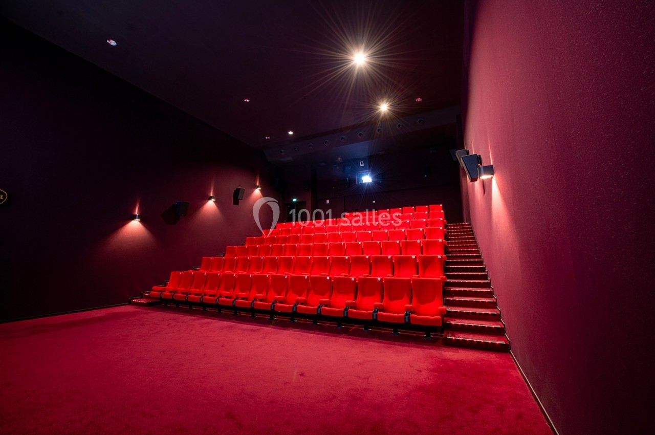 Salle de cinéma vide avec rangées de sièges rouges, éclairage tamisé et écran éteint en arrière-plan.