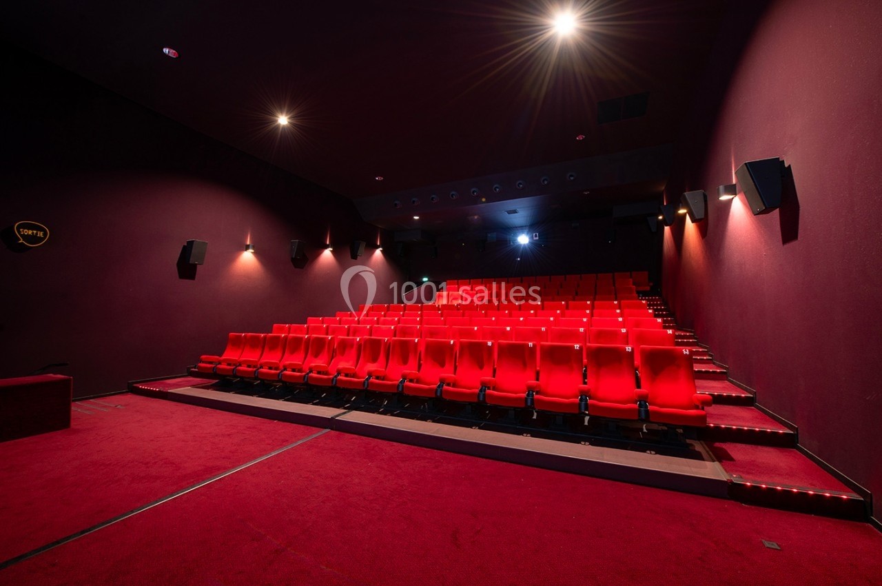 Salle de cinéma vide avec des rangées de sièges rouges, éclairée par des lumières tamisées.