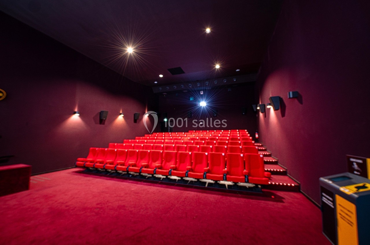 Salle de cinéma vide avec rangées de sièges rouges, éclairage tamisé et projecteur allumé.