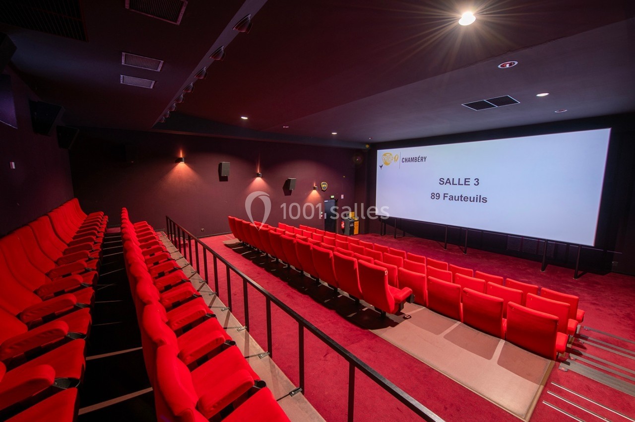 Salle de cinéma vide avec des sièges rouges, écran blanc affichant ’Salle 3, 89 fauteuils’ et éclairage tamisé.