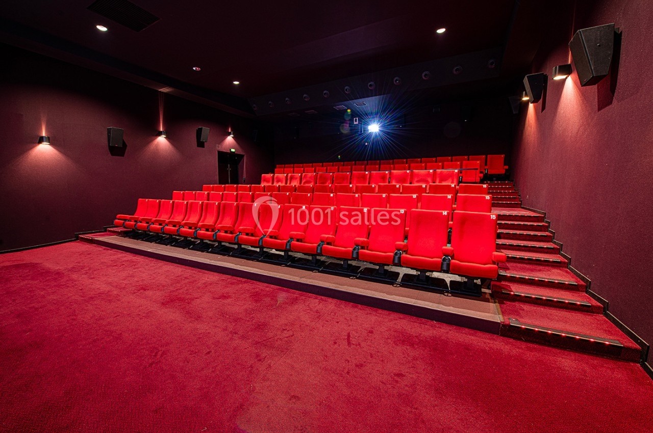 Salle de cinéma vide avec des rangées de sièges rouges et un projecteur allumé au fond.