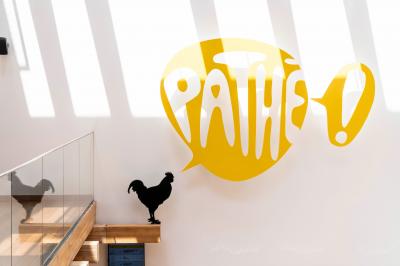 Silhouette d'un coq noir sur une rambarde en bois devant un mur blanc avec le logo jaune ’Pathé !’ éclairé par la lumière.