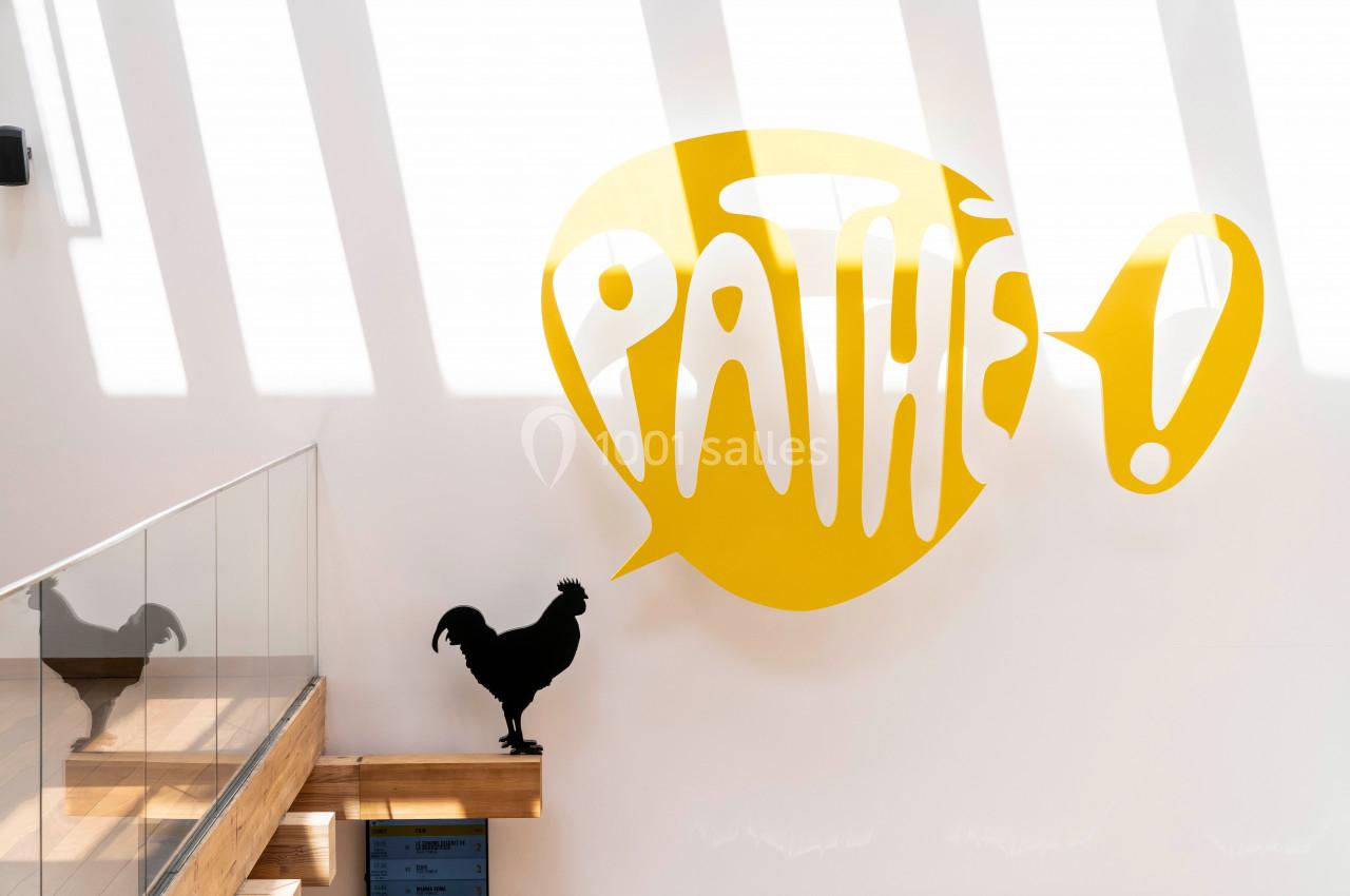 Silhouette d'un coq noir sur une rambarde en bois devant un mur blanc avec le logo jaune ’Pathé !’ éclairé par la lumière.