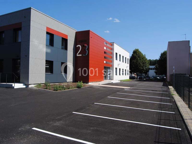 Bâtiment moderne avec façade grise et rouge, parking vide au premier plan sous un ciel dégagé.