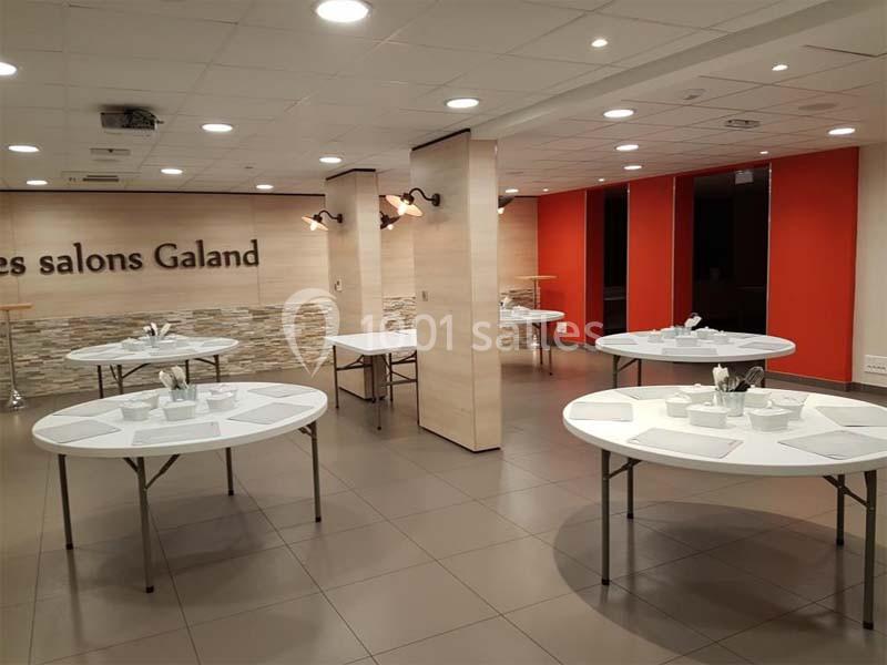 Salle lumineuse avec tables rondes dressées, murs clairs, panneaux rouges et inscription ’les salons Galand’.