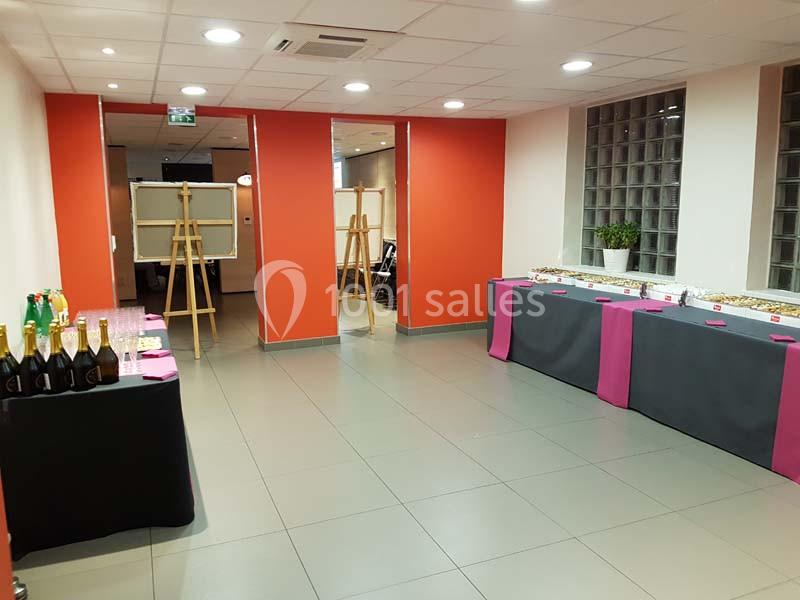 Salle lumineuse avec tables dressées pour un buffet, comprenant boissons et plats, murs rouges et baies vitrées.