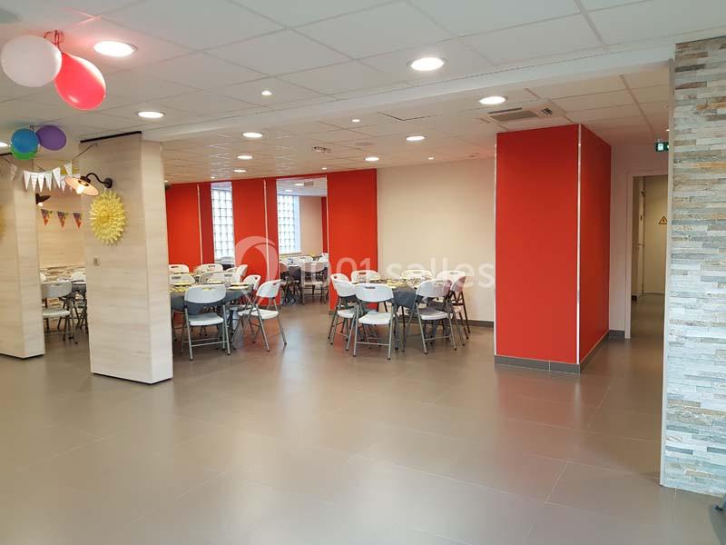 Salle lumineuse avec tables et chaises disposées pour un événement, décorée de ballons et guirlandes colorées.