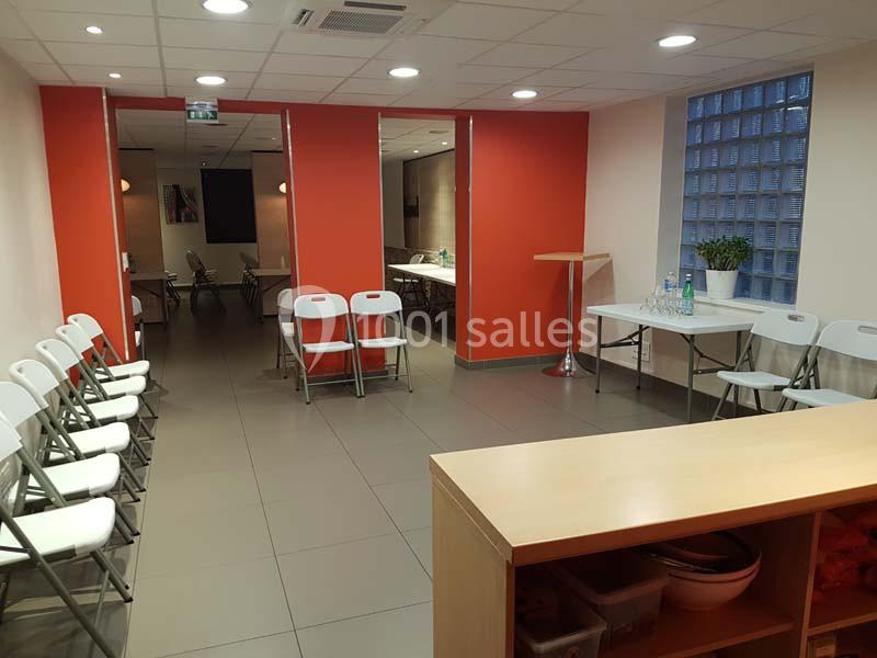 Salle lumineuse avec murs rouges, chaises pliantes alignées, tables avec bouteilles d'eau et plante décorative.