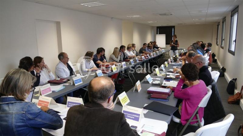 Des personnes assises autour de longues tables en réunion dans une salle lumineuse avec une présentatrice debout.