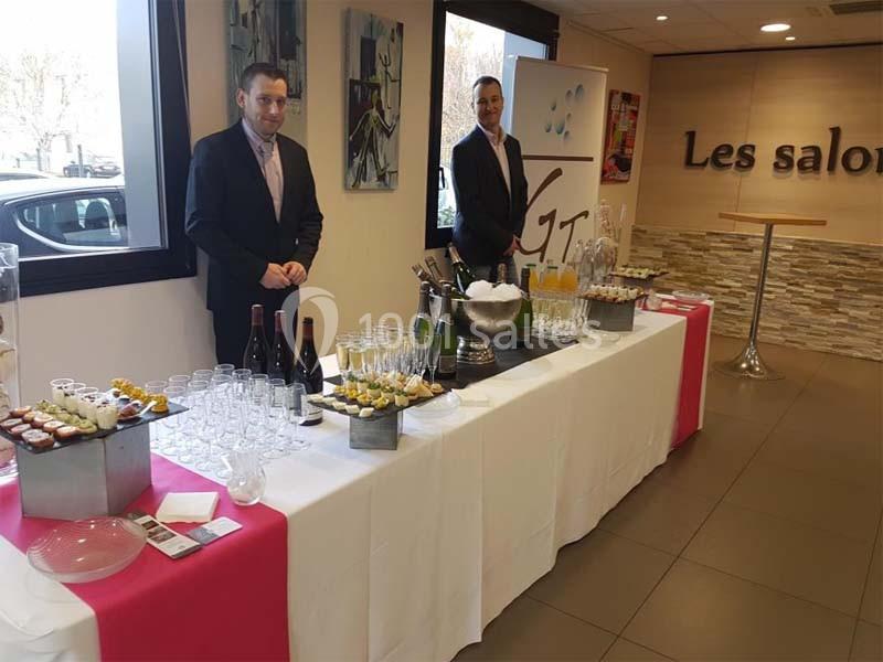 Table de réception avec boissons, verrines et desserts, deux serveurs en costume à côté, dans une salle lumineuse.