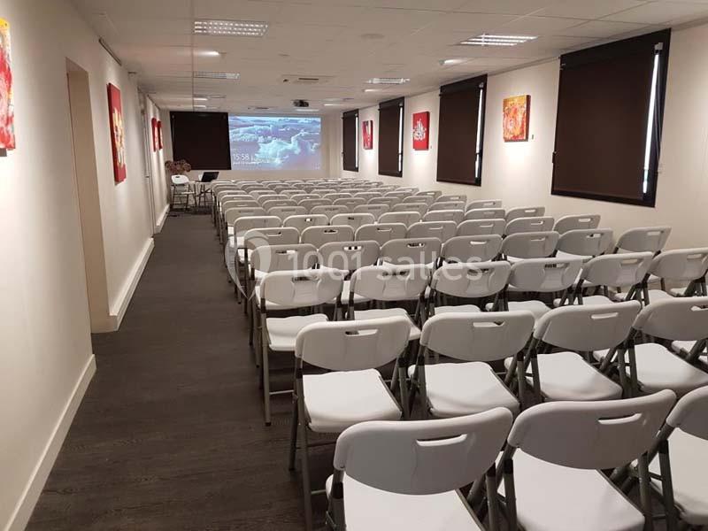 Salle de conférence avec des rangées de chaises blanches alignées face à un écran de projection.