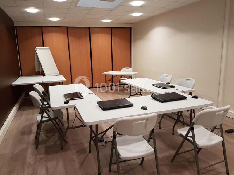 Salle de réunion équipée de tables, chaises pliantes, ordinateurs portables et un tableau papier.