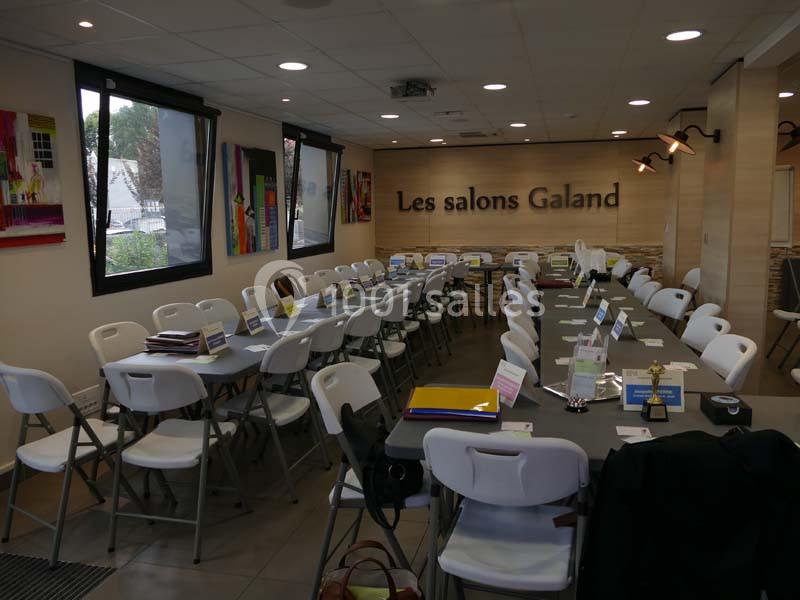 Salle de réunion aménagée avec de longues tables, des chaises blanches et des dossiers posés, éclairée par des fenêtres.