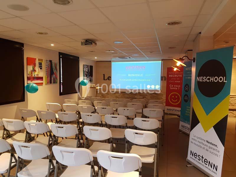 Salle de formation avec des chaises blanches alignées, un écran de projection allumé et des affiches promotionnelles.