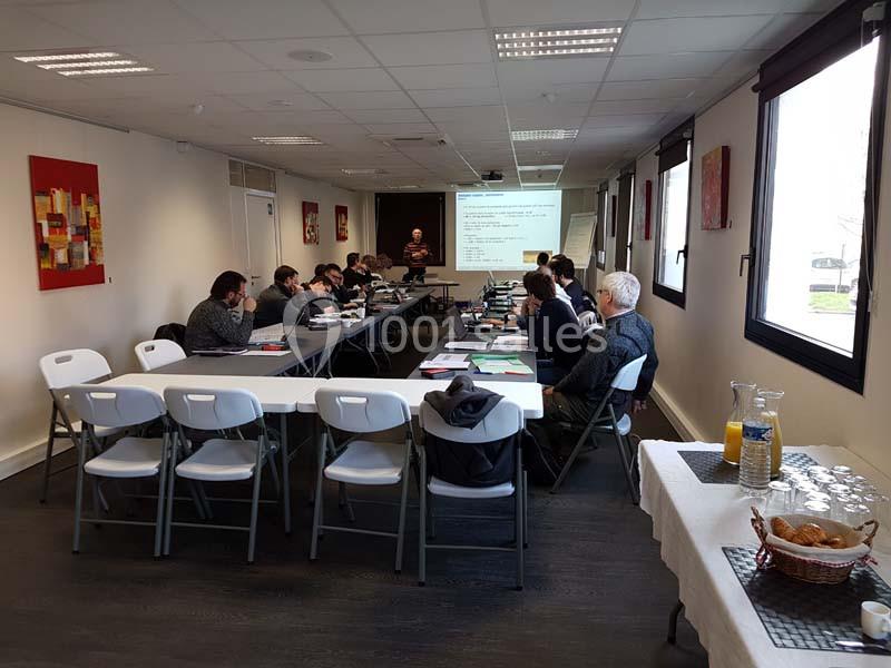 Des personnes assises autour de tables en U assistent à une présentation dans une salle lumineuse.