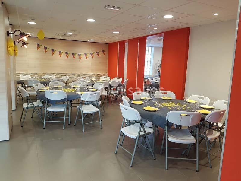 Salle décorée avec des tables rondes dressées, nappes grises, chaises blanches et guirlandes colorées accrochées au mur.