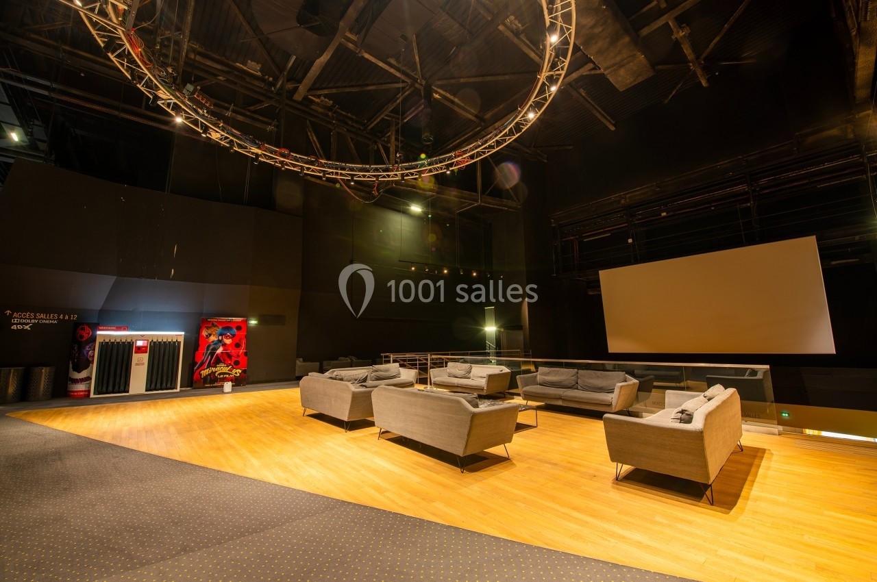 Salle avec des canapés gris, écran blanc, éclairage tamisé et structure métallique suspendue au plafond.