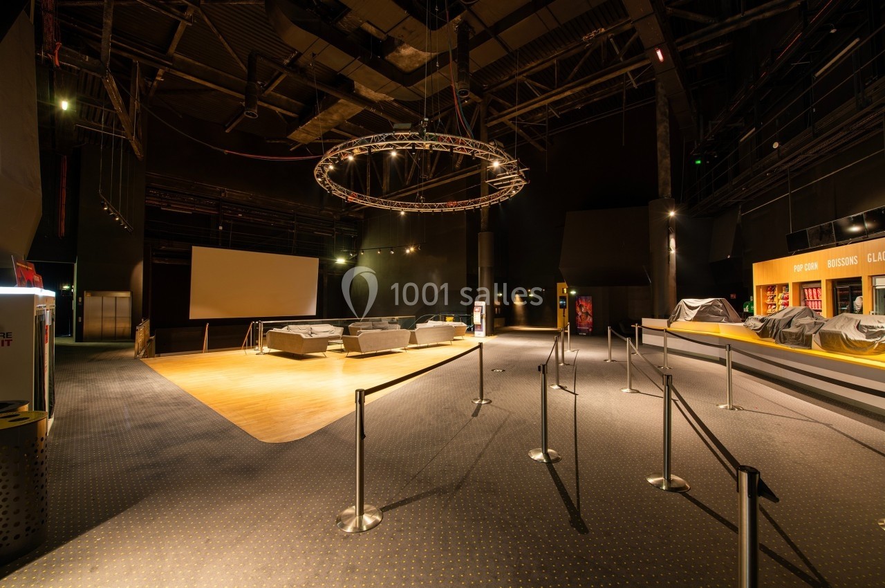 Salle sombre avec un écran géant, des sièges disposés sur une estrade et des barrières délimitant un espace.