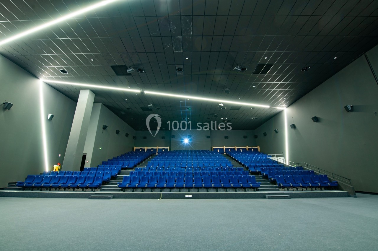 Salle de cinéma vide avec des rangées de sièges bleus et un écran éteint, éclairée par des lumières au plafond.