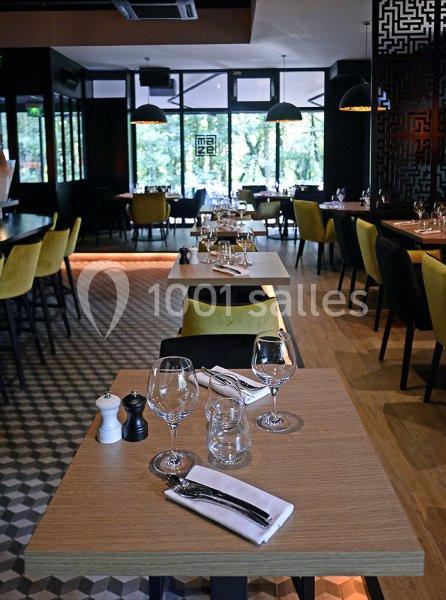 Salle de restaurant moderne avec tables dressées, chaises jaunes et grandes baies vitrées donnant sur un espace arboré.