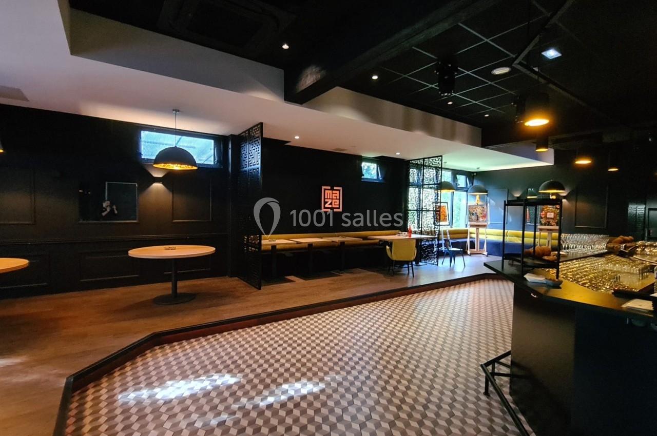 Salle moderne avec tables, banquettes, bar et éclairage tamisé, décorée de panneaux noirs et motifs géométriques au sol.