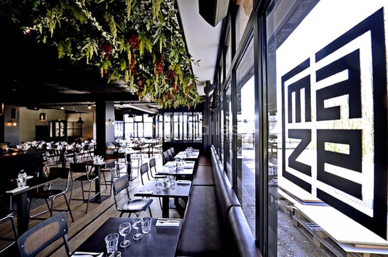 Salle de restaurant moderne avec grandes baies vitrées, tables dressées et décoration végétale suspendue.