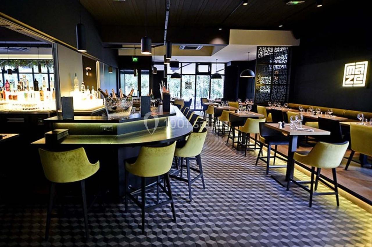 Intérieur d'un restaurant moderne avec bar central, tabourets jaunes, tables dressées et éclairage tamisé.