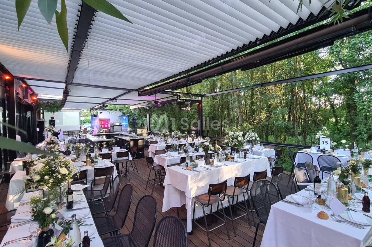 Salle extérieure décorée pour un événement, avec tables dressées, nappes blanches et arrangements floraux, entourée de…