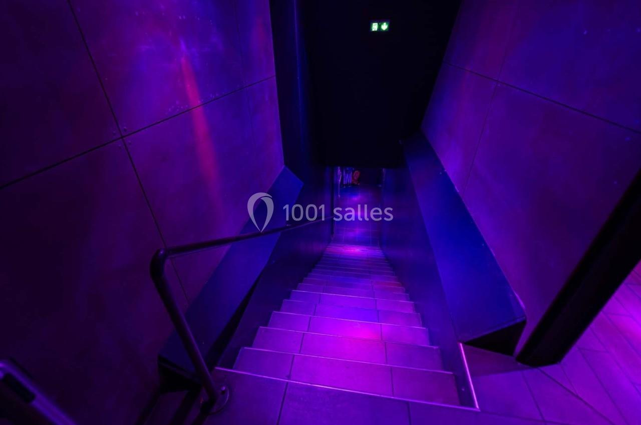 Escalier descendant dans un couloir sombre éclairé par des lumières violettes.