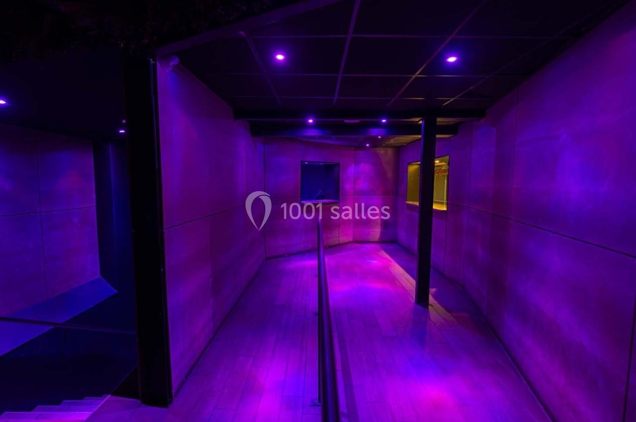 Couloir sombre éclairé par des lumières violettes, avec un sol en bois et des murs lisses.
