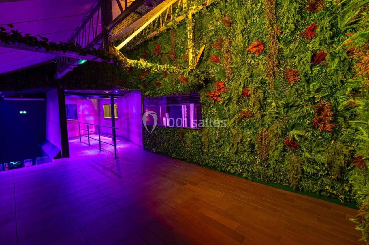 Mur végétal éclairé par des lumières colorées dans un espace intérieur avec sol en bois et structure métallique visible.